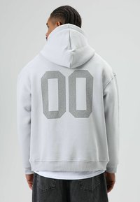 Lichtgrijze oversized hoodie van zacht materiaal, met een grote zwarte polkadot "00"-print op de achterkant en een capuchon met trekkoord.