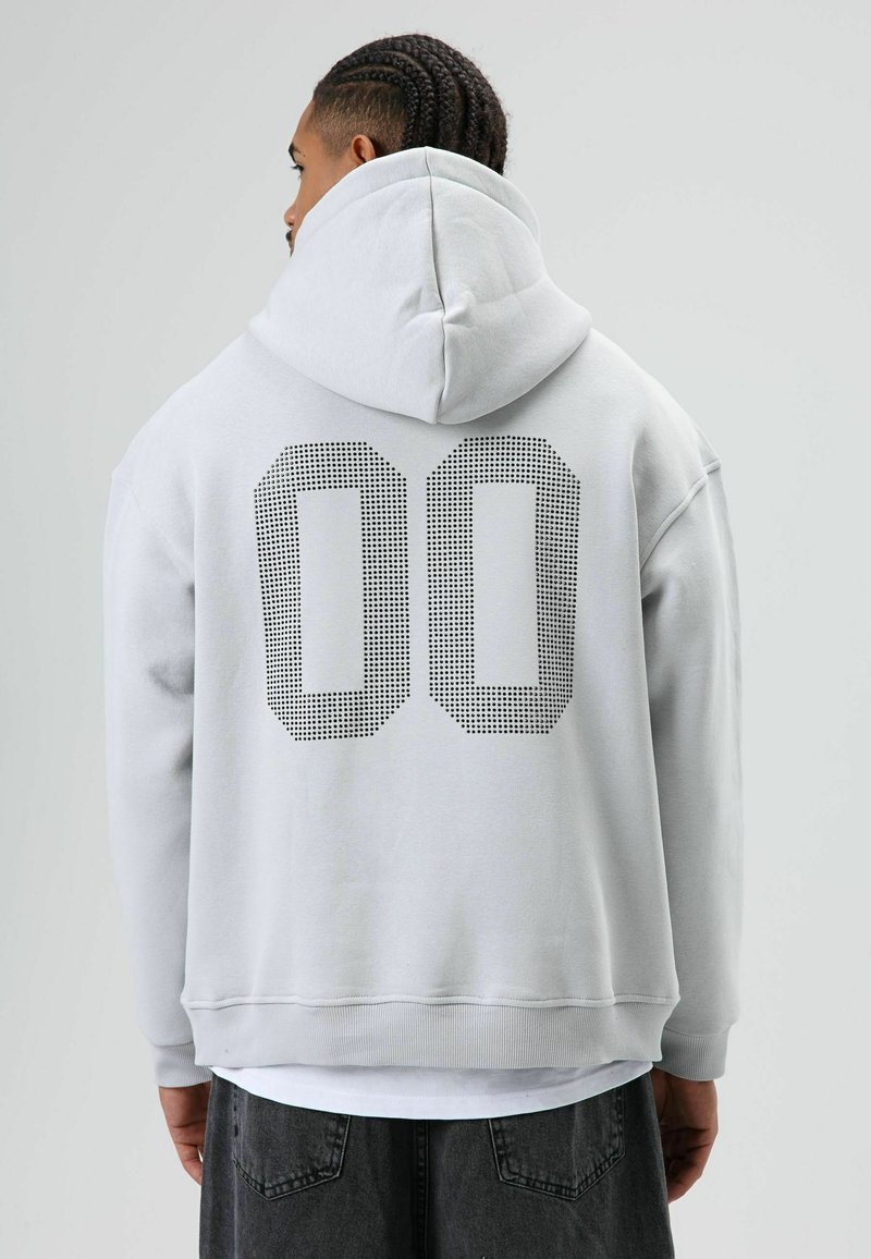 Lichtgrijze oversized hoodie van zacht materiaal, met een grote zwarte polkadot "00"-print op de achterkant en een capuchon met trekkoord.
