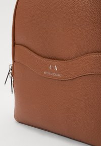 Mochila de cuero marrón texturizado con un bolsillo delantero curvado, herrajes de acero inoxidable y el logo en relieve "A/X ARMANI EXCHANGE."
