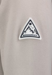 Grijze stof met een driehoekige patch met een bergontwerp, zwarte accenten, en de tekst "BERGNARRISCH" gecentreerd onderaan.
