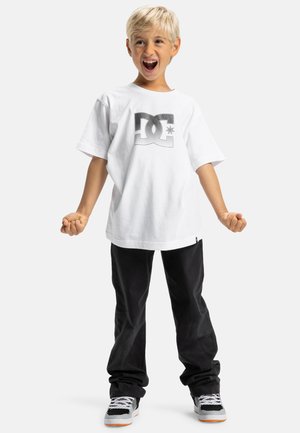 Witte katoenen t-shirt met een gedurfde zwarte DC-logo, gecombineerd met zwarte broek en grijs/zwarte sneakers met oranje accenten.