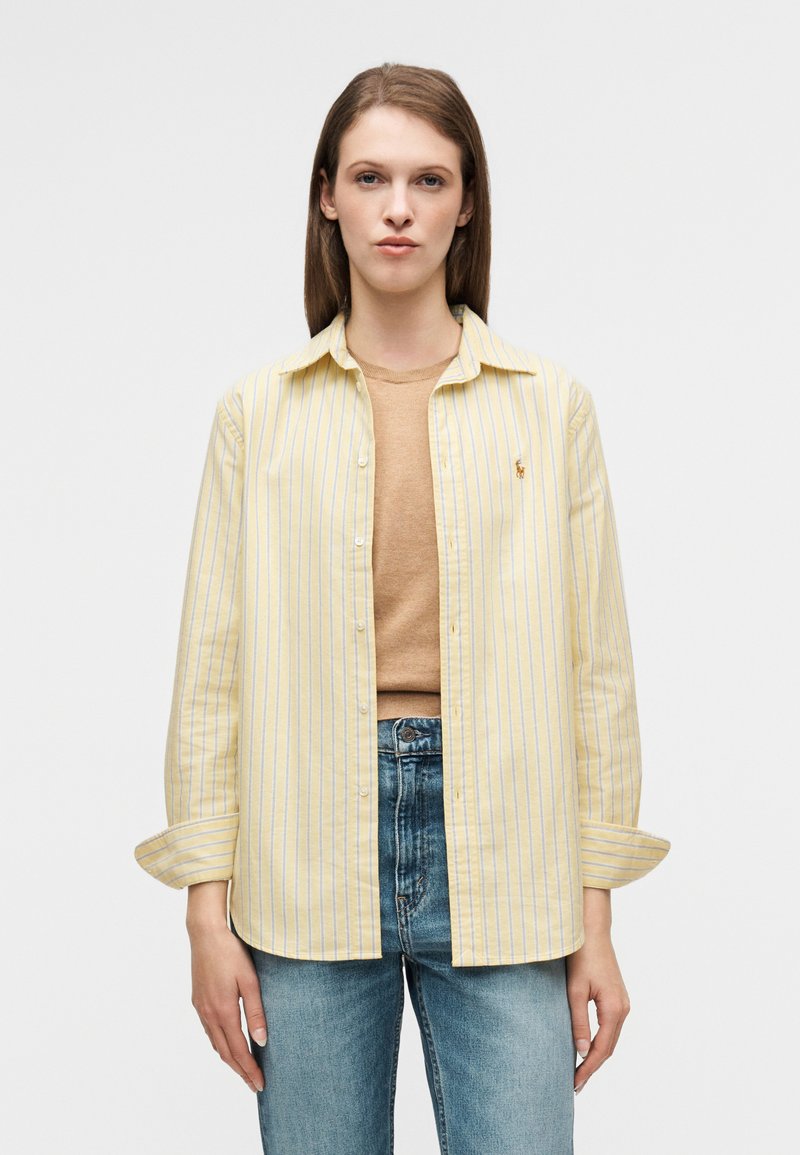 Chemise à rayures jaunes et blanches en tissu léger, avec un logo sur la poitrine et des manches retroussées. Portée par-dessus un pull beige et un jean bleu.