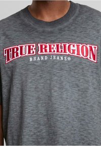 Šedé tričko s krátkým rukávem, na kterém je vyšitý výrazný červený nápis "TRUE RELIGION" a menší "BRAND JEANS" pod ním, texturovaná látka.