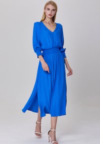 Robe bleue longue avec un décolleté en V, taille froncée et manches longues. Tissu doux et fluide avec des fentes sur les côtés. Portée avec des escarpins blancs.