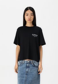 Camiseta de algodón negra con mangas cortas, escote redondo y detalle de logo en la parte frontal. Combinada con una falda de mezclilla.
