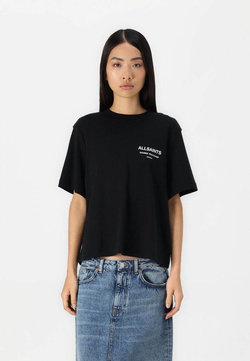 Camiseta de algodón negra con mangas cortas, escote redondo y detalle de logo en la parte frontal. Combinada con una falda de mezclilla.