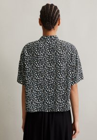 Kurzärmliges, tailliertes schwarzes Shirt mit einem weißen und hellblauen Musterdesign, das verschiedene kleine Formen und Symbole zeigt. Glatte Textur.