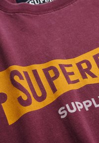 Bordeaux katoenen t-shirt met een gedurfde gele en witte grafische print met de tekst "SUPER" en "SUPPLY." Zachte textuur, ronde hals.