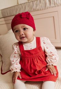 Robe en velours côtelé rouge avec ourlet à volants, chemise blanche à manches longues avec motif de cerises, et bonnet en tricot rouge avec détail de nœud.