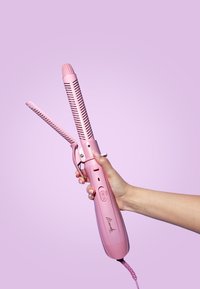 Fer à friser rose avec une prise en main texturée, un cylindre perforé et un interrupteur marche/arrêt. Main tenant l'appareil, avec un cordon d'alimentation raccordé.