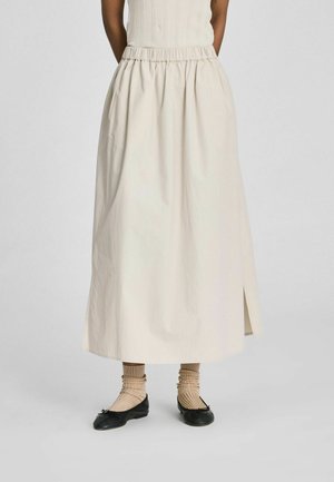 Frau trägt einen langen cremefarbenen Rock mit seitlichem Schlitz, kombiniert mit beigen Socken und schwarzen Ballettschuhen, steht vor einem schlichten Hintergrund.