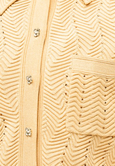 Μπεζ πλεκτό ύφασμα με σχέδιο ζικ-ζακ, μικρές μεταλλικές τρουκς, εμπρόσθια τσέπη και κουμπιά σε σχήμα λουλουδιού με μαργαριταρένια κέντρα.