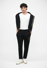 Zwart sweatshirt met logo op de mouw, gecombineerd met zwarte sweatpants met gouden print. Witte T-shirt eronder en witte sneakers.