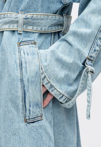 Mão no bolso de uma jaqueta de ganga azul claro com cinto e costura detalhada na manga e no bolso.