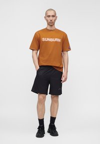 Homme debout, face vers l'avant, portant un T-shirt marron avec le texte "SUNBURN", short noir, chaussettes noires et baskets noires sur un fond uni.