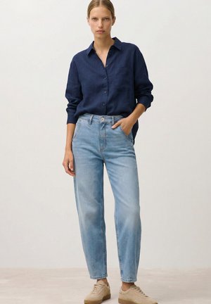 Woman standing in casual outfit, wearing a navy button-up shirt, light blue jeans, and beige sneakers, one hand in pocket against plain background.