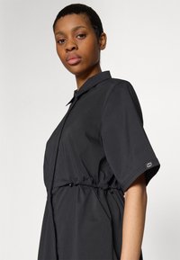 Abito nero a maniche corte con collo e vita arricciata. Presenta un tessuto morbido e un design minimalista, con un logo discreto sulla manica.