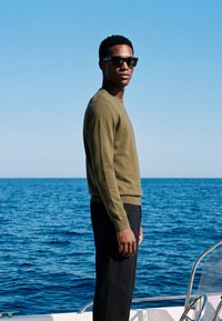 Homme portant des lunettes de soleil, un pull vert olive et un pantalon noir, debout sur un bateau avec une mer bleue et un ciel dégagé en arrière-plan.