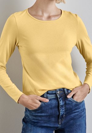 Żółta koszula z długim rękawem i okrągłym dekoltem, zestawiona z niebieskimi dżinsami. Tkanina wydaje się gładka i elastyczna.