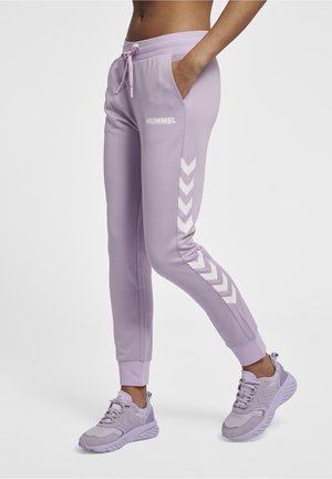 Woman wearing light purple Hummel joggers with white chevron stripes and matching sneakers on a white background.