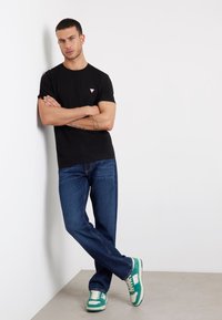 Juodas medvilninis marškinėliai su mažu raudonu trikampio logotipu, derinami su mėlynomis džinsinėmis kelnes ir žaliais sportiniais batais su baltais akcentais bei tekstūruotu padu.