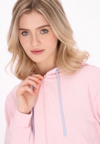 Helle rosa Kapuzenjacke mit Reißverschluss vorne, kontrastierenden hellblauen Kordeln und gerippten Bündchen. Weiches Material mit glatter Textur.