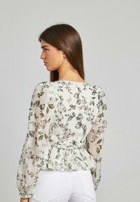 Blusa floreale in tessuto leggero e trasparente con maniche a palloncino, orlo peplum e dettaglio con fiocco sul retro. Predominante color crema con motivi verdi e rosa.