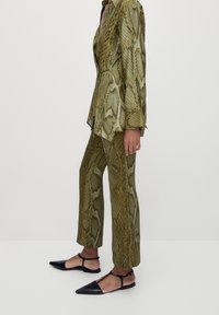 Costume vert à imprimé serpent avec un blazer structuré et un pantalon droit. Chaussures plates noires à bout pointu et bride de cheville. Motif de tissu texturé.