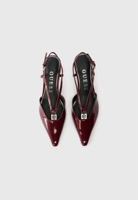 Ζεύγος γυαλιστερών μπορντώ γόβων με μυτερή μύτη και λουριά στον αστράγαλο, με λεπτομέρεια από ασημένιο κούμπωμα και το λογότυπο "GUESS" στο μαύρο σόλο.