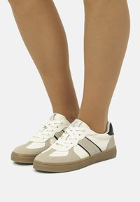 Sneaker mit einer Mischung aus Wildleder und Leder in Beige und Weiß, mit schwarzen Akzenten und klassischen weißen Schnürsenkeln. Robuste Gummisohle.