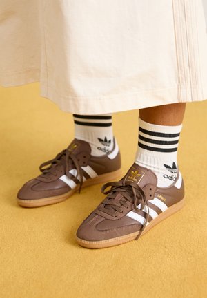 Osoba nosząca brązowe tenisówki Adidas Samba oraz białe skarpetki Adidas z czarnymi paskami, stojąca na musztardowo-żółtej podłodze, widoczna biała spódnica.