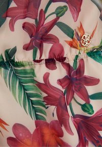 Blomsterpræget stof med store lyserøde og orange blomster og grønne blade, pyntet med et lille metallisk logo på sidesømmen.