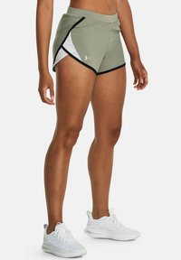 Mujer vestida con shorts de running de color verde olivo de Under Armour con ribetes negros y zapatillas blancas, de pie contra un fondo liso.