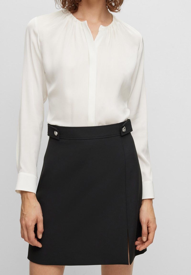 Biała, długa bluzka z długim rękawem i marszczonym dekoltem. W połączeniu z czarną, dopasowaną spódnicą z guzikami i bocznym rozporkiem.