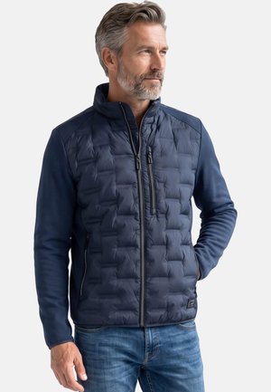 Mittelalter Mann mit grauem Haar und Bart, der eine dunkelblaue gesteppte Jacke mit Reißverschlusstaschen und blaue Jeans trägt, steht mit einer Hand in der Tasche.