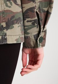 Main reposant à côté du corps, portant une veste camouflage verte et marron avec un poignet boutonné et un pantalon noir sur un fond clair.