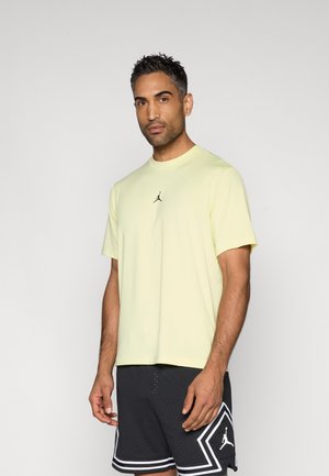 Világos sárga rövid ujjú póló kerek nyakkivágással és egy kis fekete Jordan logóval a mellkasán, fekete mintás rövidnadrággal párosítva.