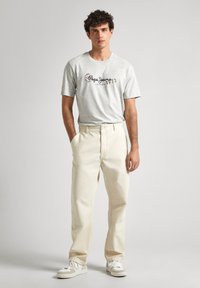 Camiseta de algodón gris con un estampado gráfico, combinada con pantalones de algodón color crema, complementada con zapatillas blancas. Modelo de pie.