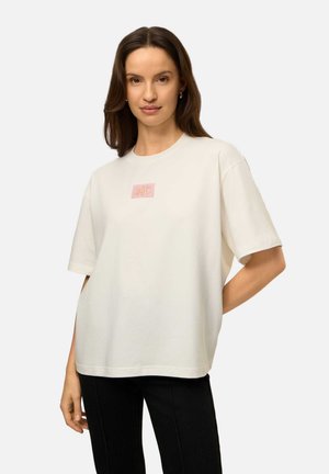 Vrouw met bruin haar die een los wit t-shirt draagt met een klein roze logo in het midden en zwarte broek, staand met één hand achter haar rug.