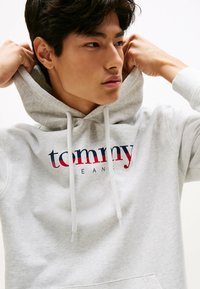 Hoodie gris en matière polaire, avec un grand logo "tommy" en rouge et bleu marine, équipé d'une capuche à cordon de serrage et d'une poche avant.