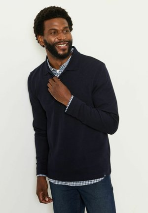 Smilende mand med skæg iført navy sweater over blå og hvide ternet skjorte, stående mod hvid baggrund.