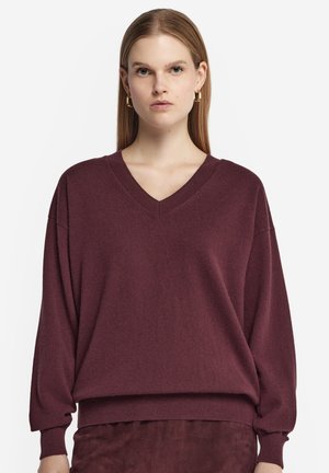Femme portant un pull ample à manches longues bordeaux à col en V et une jupe assortie, avec des cheveux lisses et des boucles d'oreilles en anneau doré.