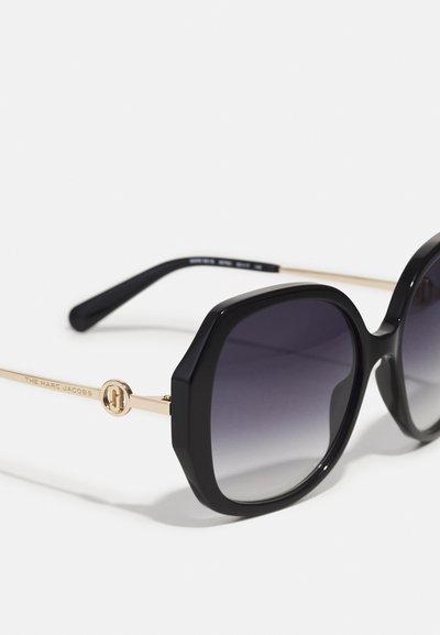 Schwarze oversized Sonnenbrille mit goldenen Akzenten, ausgestattet mit einem klobigen Kunststoffrahmen und einem verlaufenden dunklen Glas, beschriftet mit "THE MARC JACOBS."