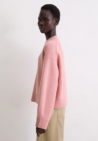 Hellrosa, gestrickter Pullover mit gerippten Bündchen und Saum, der eine lockere Passform und einen Rundhalsausschnitt bietet. Das Modell wird im Seitenprofil dargestellt.