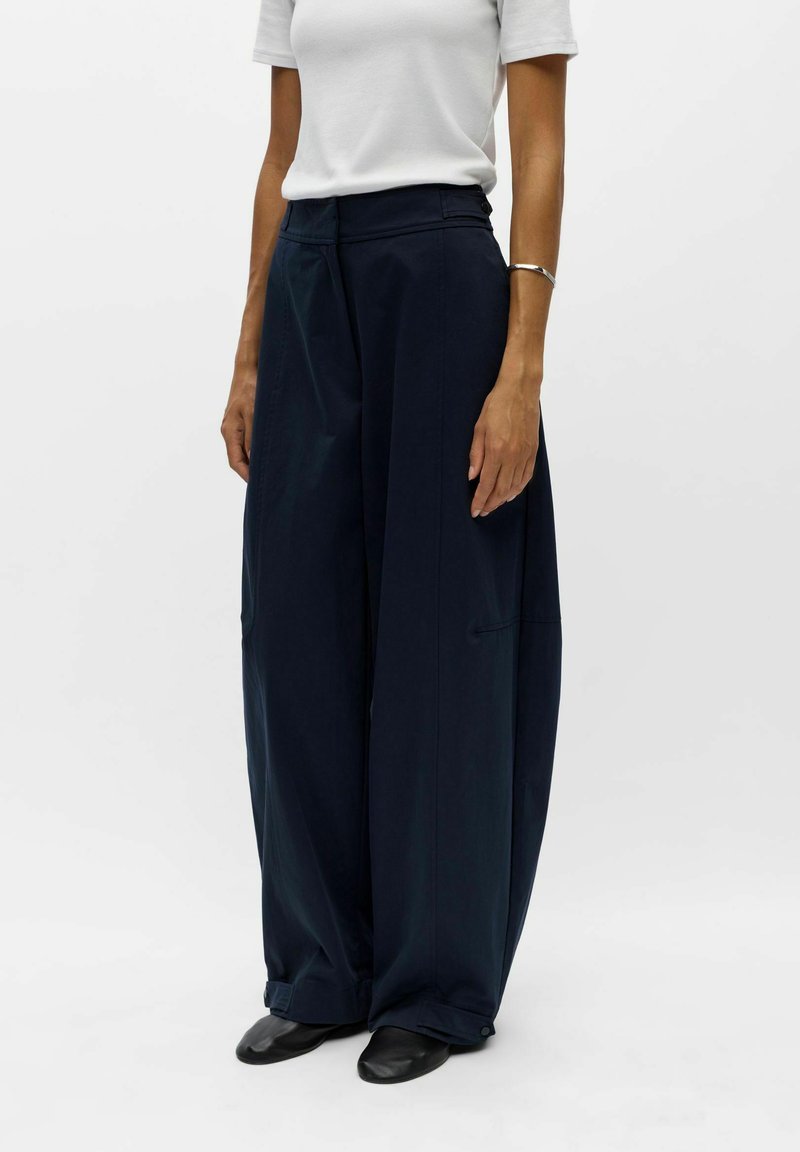 Personne portant un pantalon large bleu marine foncé, un haut blanc à manches courtes, des chaussures noires et un bracelet argenté au poignet gauche.