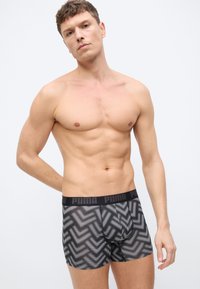 Homme torse nu portant un boxer Puma à motifs noirs et blancs, debout devant un fond clair uni avec une main derrière le dos.