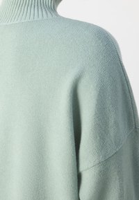 Pull-over en tricot de couleur vert menthe doux, avec un col montant à côtes et une coupe décontractée, dotée d'un tricot texturé. Souligné par des épaules tombantes.