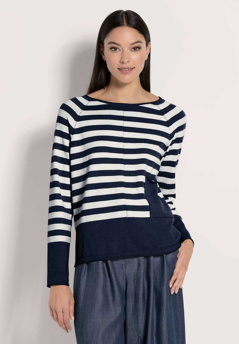 Frau mit langen, dunklen Haaren, die einen navy- und weißgestreiften Pullover und lockere blaue Hosen trägt, steht vor einem schlichten hellen Hintergrund.