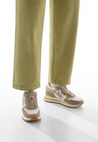 Beżowe i białe sneakersy z grubą podeszwą noszone z jasnymi spodniami khaki, stojące na białej powierzchni z widocznymi cieniami.