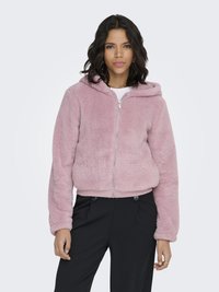 Woman wearing a light pink zip-up fuzzy hoodie with a hood, paired with high-waisted black trousers against a plain background.